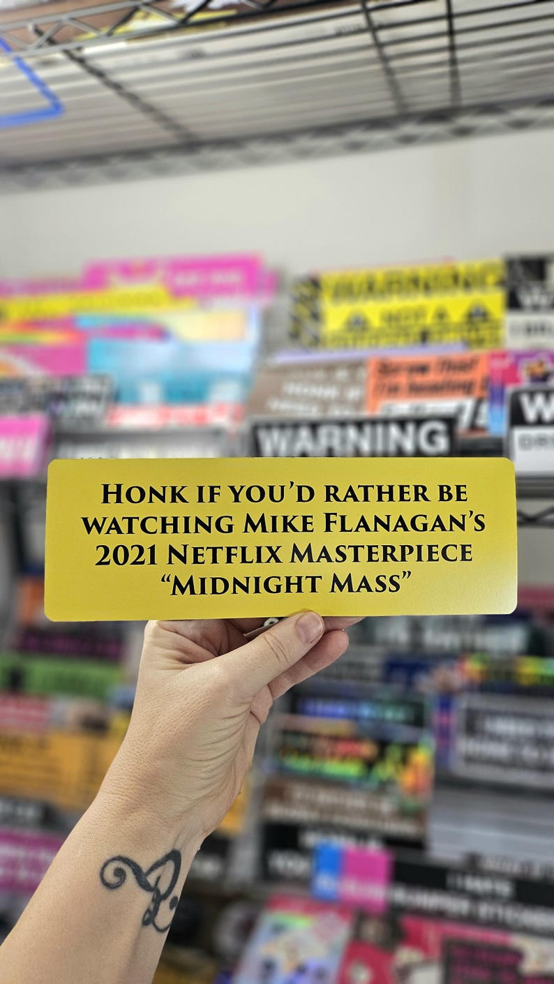 Peut inclure: Un panneau de couleur or avec du texte noir qui dit "HONK IF YOU'D RATHER BE WATCHING MIKE FLANAGAN'S 2021 NETFLIX MASTERPIECE "MIDNIGHT MASS""