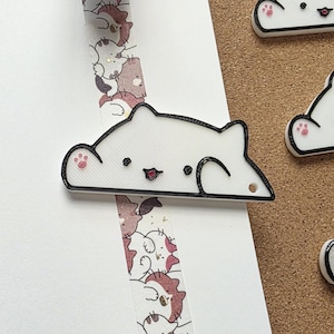 May include: White cat-shaped decorative pins with black outlines and pink paw prints. The pins have a small hole for attaching to items. A roll of decorative tape with cat illustrations is also visible.