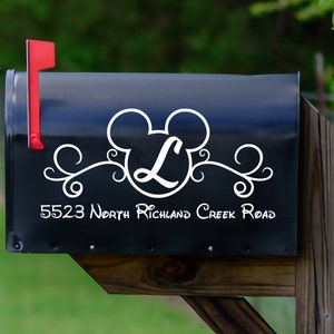 Puede incluir: Buzón negro con una bandera roja. Presenta un diseño blanco inspirado en Mickey Mouse con la letra "L" y adornos decorativos. La dirección "5523 North Richland Creek Road" también se muestra en blanco.
