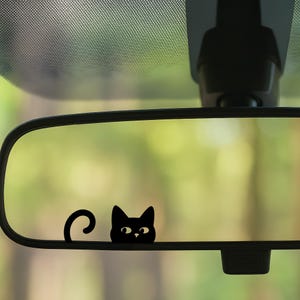 May include: Black cat decal on a car's rearview mirror. The cat has a curious expression with large eyes and a curved tail. The background is a blurred view of green foliage.
