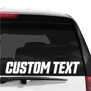 May include: White vinyl decal with the words "CUSTOM TEXT" in a bold, slanted font, applied to the rear window of a vehicle. The decal is positioned above the rear windscreen wiper.