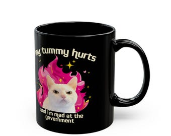 May include: Black coffee mug with a white cat in a pink flame design. The text on the mug reads "my tummy hurts and I'm mad at the government."