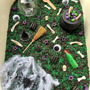 May include: A sensory bin filled with green and brown rice, featuring a clear glass bottle with a cork, a black plastic cauldron, a miniature broom, a green finger puppet, a black spider on white webbing, googly eyes, and small plastic bones.