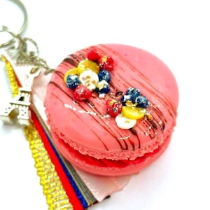 May include: A pink macaron keychain with a silver Eiffel Tower charm and a decorative ribbon. The macaron is topped with berries and chocolate drizzle. The keychain is on a white background.