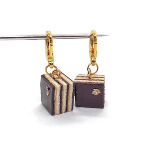 Peut inclure: Boucles d'oreilles dorées avec des breloques miniatures en forme de gâteau à plusieurs couches. Les breloques sont brun foncé avec des couches brun clair et crème, et une petite décoration dorée. Les boucles d'oreilles sont présentées sur fond blanc.