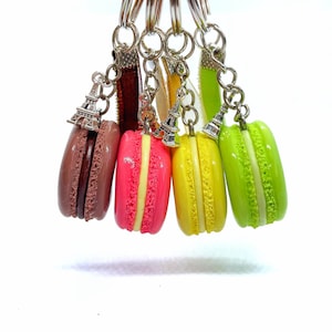 Könnte beinhalten: Eine Sammlung bunter Macaron-Schlüsselanhänger. Jeder Schlüsselanhänger hat einen Miniatur-Eiffelturm-Anhänger und ein Macaron in Braun-, Rosa-, Gelb- und Grüntönen. Die Macarons haben eine cremefarbene Füllung.