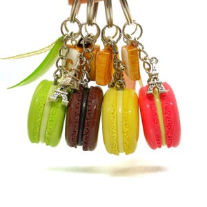 Könnte beinhalten: Eine Sammlung bunter Macaron-Schlüsselanhänger in Grün, Braun, Gelb und Pink. Jeder Schlüsselanhänger hat einen Miniatur-Eiffelturm-Anhänger und eine silberne Kette. Die Macarons haben eine cremefarbene Füllung.
