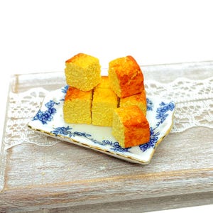 Peut inclure: Une petite assiette rectangulaire avec un motif floral bleu présente plusieurs carrés de gâteau jaune doré. Le gâteau a un aspect texturé et est disposé sur une assiette blanche et dorée. L'assiette repose sur une surface en bois.