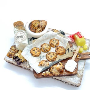 Peut inclure: Une scène de pâtisserie miniature avec des cookies aux pépites de chocolat, un pot de farine et un petit bol de cookies. Les cookies sont sur une planche à découper en bois avec une petite spatule. Une serviette à carreaux rouges et blancs est également présente.