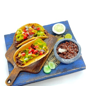 Peut inclure: Miniatures de tacos garnis, présentés sur une planche en bois posée sur une surface bleue. Un petit bol de chili et des quartiers de citron vert sont également présents. Les tacos ont des coquilles jaunes, de la laitue verte, des tomates rouges et une garniture brune.