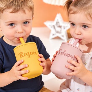 Puede incluir: Dos ni&ntilde;os bebiendo de vasos personalizados. Un vaso es de color amarillo mostaza con el nombre "Regan" impreso, y el otro es de color rosa empolvado con el nombre "Solomon". Ambos vasos tienen pajitas a juego.