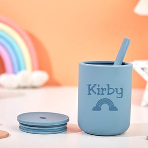 Puede incluir: Una taza de silicona azul claro con tapa y pajita a juego. La taza est&aacute; personalizada con el nombre "Kirby" y un gr&aacute;fico de arco&iacute;ris. Un arco&iacute;ris y una decoraci&oacute;n en forma de estrella est&aacute;n en el fondo.