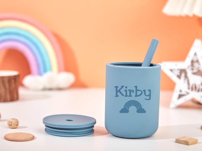 Puede incluir: Una taza de silicona azul claro con tapa y pajita a juego. La taza est&aacute; personalizada con el nombre "Kirby" y un gr&aacute;fico de arco&iacute;ris. Un arco&iacute;ris y una decoraci&oacute;n en forma de estrella est&aacute;n en el fondo.