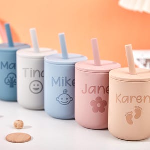 Puede incluir: Cinco vasos personalizados de silicona con pajitas en azul, blanco, azul claro, rosa y melocot&oacute;n. Cada vaso presenta un nombre y un icono o dise&ntilde;o &uacute;nico. Los nombres son "Ma", "Tina", "Mike", "Jane" y "Karen".