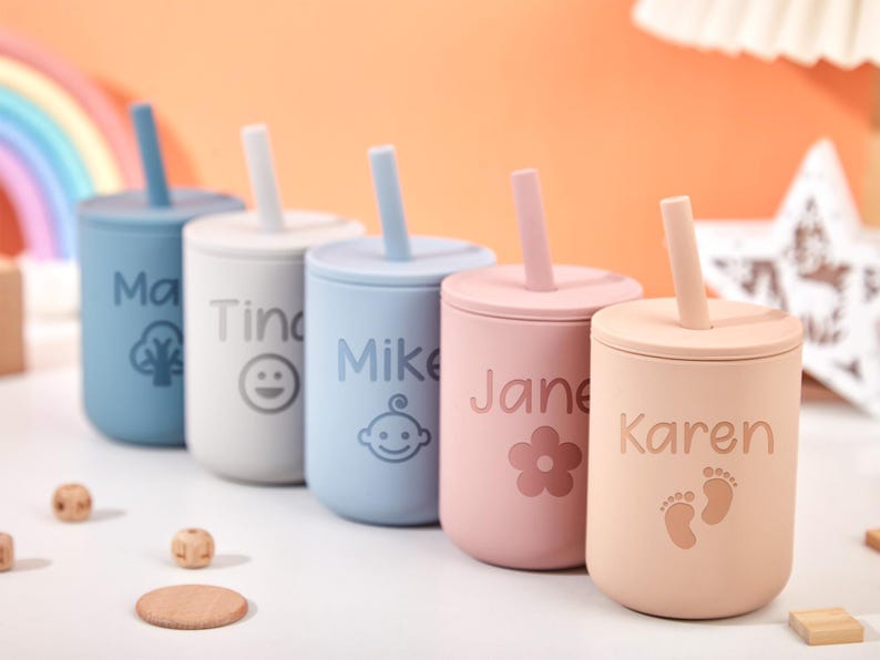 Puede incluir: Cinco vasos personalizados de silicona con pajitas en azul, blanco, azul claro, rosa y melocot&oacute;n. Cada vaso presenta un nombre y un icono o dise&ntilde;o &uacute;nico. Los nombres son "Ma", "Tina", "Mike", "Jane" y "Karen".