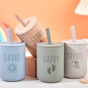 Puede incluir: Cuatro vasos con boquilla personalizados para beb&eacute;s en colores pastel: azul, beige, gris y verde. Cada vaso tiene un nombre: David, Frank, Harry y Travis. El vaso azul tiene un sol sonriente, el vaso Harry tiene huellas y el vaso Travis tiene una abeja.