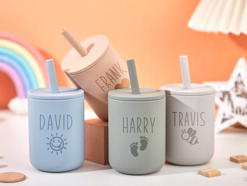 Puede incluir: Cuatro vasos con boquilla personalizados para beb&eacute;s en colores pastel: azul, beige, gris y verde. Cada vaso tiene un nombre: David, Frank, Harry y Travis. El vaso azul tiene un sol sonriente, el vaso Harry tiene huellas y el vaso Travis tiene una abeja.