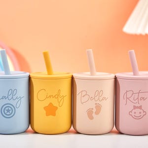 Puede incluir: Cuatro vasos coloridos para beb&eacute;s con tapas y pajitas. Cada vaso es de un color diferente: azul, amarillo, beige y rosa. Los vasos est&aacute;n personalizados con nombres y s&iacute;mbolos: "Sally" con una estrella, "Cindy" con una estrella, "Bella" con huellas y "Rita" con una cara de beb&eacute;.