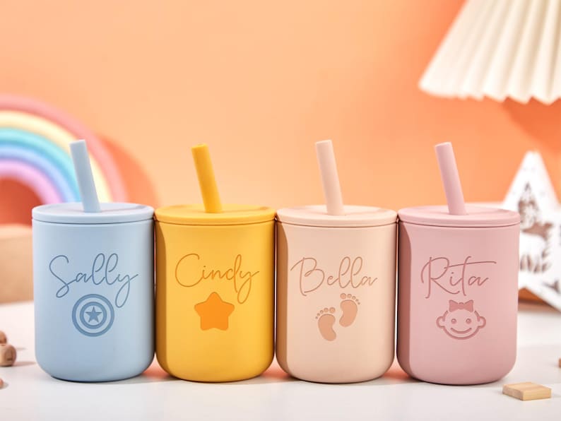 Puede incluir: Cuatro vasos coloridos para beb&eacute;s con tapas y pajitas. Cada vaso es de un color diferente: azul, amarillo, beige y rosa. Los vasos est&aacute;n personalizados con nombres y s&iacute;mbolos: "Sally" con una estrella, "Cindy" con una estrella, "Bella" con huellas y "Rita" con una cara de beb&eacute;.