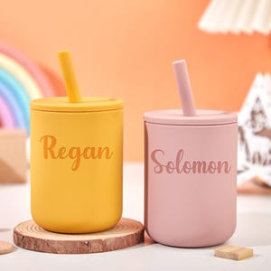 Puede incluir: Dos vasos de silicona personalizados con tapas y pajitas. Uno es amarillo con "Regan" impreso, y el otro es rosa con "Solomon" impreso. Los vasos est&aacute;n sobre posavasos de madera, con un arco&iacute;ris y una estrella de fondo.