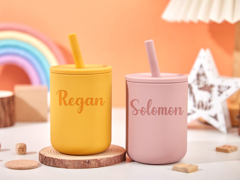 Puede incluir: Dos vasos de silicona personalizados con tapas y pajitas. Uno es amarillo con "Regan" impreso, y el otro es rosa con "Solomon" impreso. Los vasos est&aacute;n sobre posavasos de madera, con un arco&iacute;ris y una estrella de fondo.