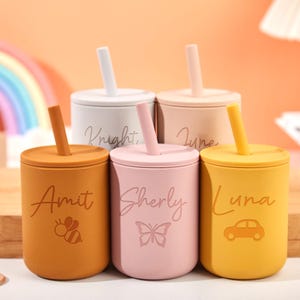 Puede incluir: Un juego de cinco tazas de silicona de colores con tapas y pajitas. Cada taza es de un color diferente: naranja, rosa, amarillo, beige y blanco. Las tazas est&aacute;n personalizadas con nombres y peque&ntilde;as ilustraciones, incluyendo una abeja, una mariposa y un coche.