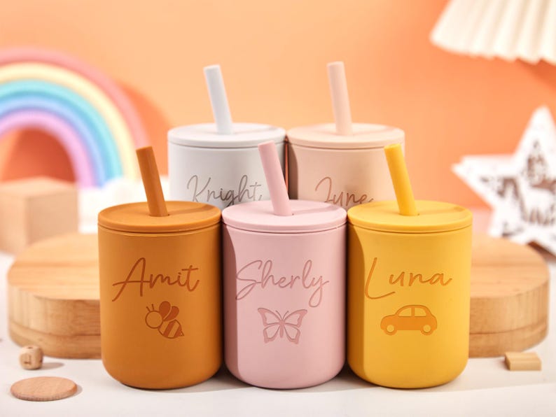 Puede incluir: Un juego de cinco tazas de silicona de colores con tapas y pajitas. Cada taza es de un color diferente: naranja, rosa, amarillo, beige y blanco. Las tazas est&aacute;n personalizadas con nombres y peque&ntilde;as ilustraciones, incluyendo una abeja, una mariposa y un coche.