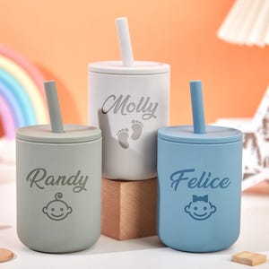 Puede incluir: Tres vasos con pajita personalizados para beb&eacute;s en colores pastel: gris claro, blanco y azul. Cada vaso tiene un nombre y un gr&aacute;fico de cara de beb&eacute;. Los vasos tienen pajitas y tapas a juego. El fondo incluye un arco&iacute;ris y bloques de madera.