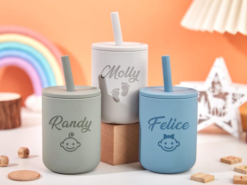 Puede incluir: Tres vasos con pajita personalizados para beb&eacute;s en colores pastel: gris claro, blanco y azul. Cada vaso tiene un nombre y un gr&aacute;fico de cara de beb&eacute;. Los vasos tienen pajitas y tapas a juego. El fondo incluye un arco&iacute;ris y bloques de madera.