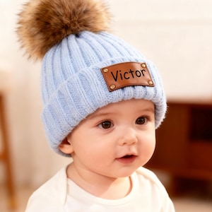 Puede incluir: Un gorro de punto azul claro con un pompón de piel sintética marrón. El sombrero presenta un parche de cuero marrón con el nombre "Victor" en letras negras. El bebé lleva un body blanco.