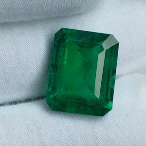 Puede incluir: Una vibrante esmeralda rectangular, que muestra un tono verde intenso. La gema se presenta sobre un fondo de tela blanca y hierba verde, resaltando su claridad y brillo. Las facetas de la esmeralda reflejan la luz, enfatizando su corte y calidad.
