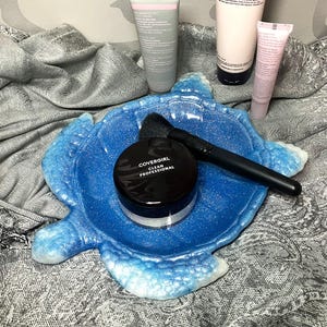 May include: A blue, turtle-shaped resin tray holds a black makeup brush, a jar of Covergirl Clean Professional powder, and several tubes of skincare products. The products include a 4-in-1 cleanser and night cream.