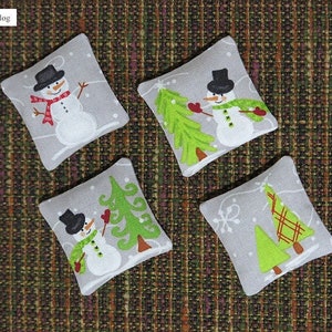May include: Four gray fabric squares with embroidered designs of snowmen and Christmas trees. The snowmen are wearing black hats and red scarves. The trees are green with brown trunks.