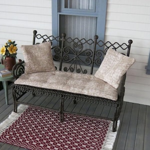 May include: Miniature wrought iron bench with two beige cushions, set on a wooden porch with a patterned rug. A small table with a flower pot and book is to the left. The bench has decorative scrollwork.
