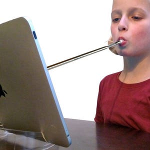 Peut inclure: Une jeune personne aux cheveux blonds en couettes utilise une longue paille argentée pour boire à partir d'une tablette posée sur un support transparent. La tablette est argentée et porte un logo Apple au dos.