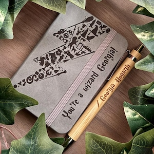 May include: A grey notebook featuring a lightning bolt design filled with shapes, and the text "You're a wizard Georgia!" alongside a wooden pen engraved with "Georgia Hogarth". Green leaves surround the items, set on a wooden surface.