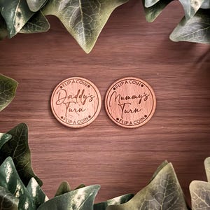 May include: Two wooden tokens with engraved text. One says "Daddy's Turn" and the other says "Mummy's Turn", both with "Flip a Coin" around the edge. The tokens are set on a wooden surface, surrounded by green leaves.