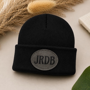 May include: A solid black knit beanie featuring a folded cuff. Centred on the cuff is an oval-shaped grey patch with the letters JRDB printed in a bold, dark font. The hat is constructed from a ribbed fabric with a consistent texture throughout.