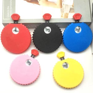 May include: Five circular measuring tapes in red, black, blue, pink, and yellow. Each tape features a textured edge, a small central window displaying numerical markings, and a red or yellow pull tab at the top for extending the tape measure.