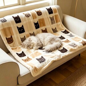 Peut inclure: Une couverture crème avec un motif répété de visages de chats de différentes couleurs, dont marron, noir et gris. Un chat duveteux gris clair est endormi sur la couverture, drapée sur un canapé beige.