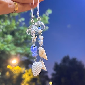 Peut inclure: Deux breloques suspendues composées de cloches en verre transparent, d'étoiles de mer blanches, de perles en forme de poisson bleu et de divers coquillages blancs. Elles sont fixées par des cordons blancs avec des fermoirs en métal argenté.