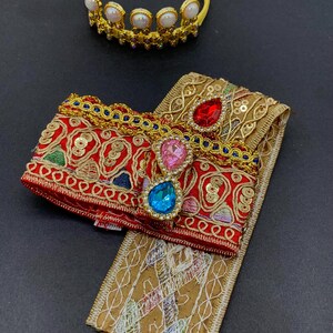 May include: A gold-colored crown with white pearl-like accents and a yellow headband. A red and gold embroidered accessory with colorful gems and a gold-colored trim. A gold-colored embroidered fabric.