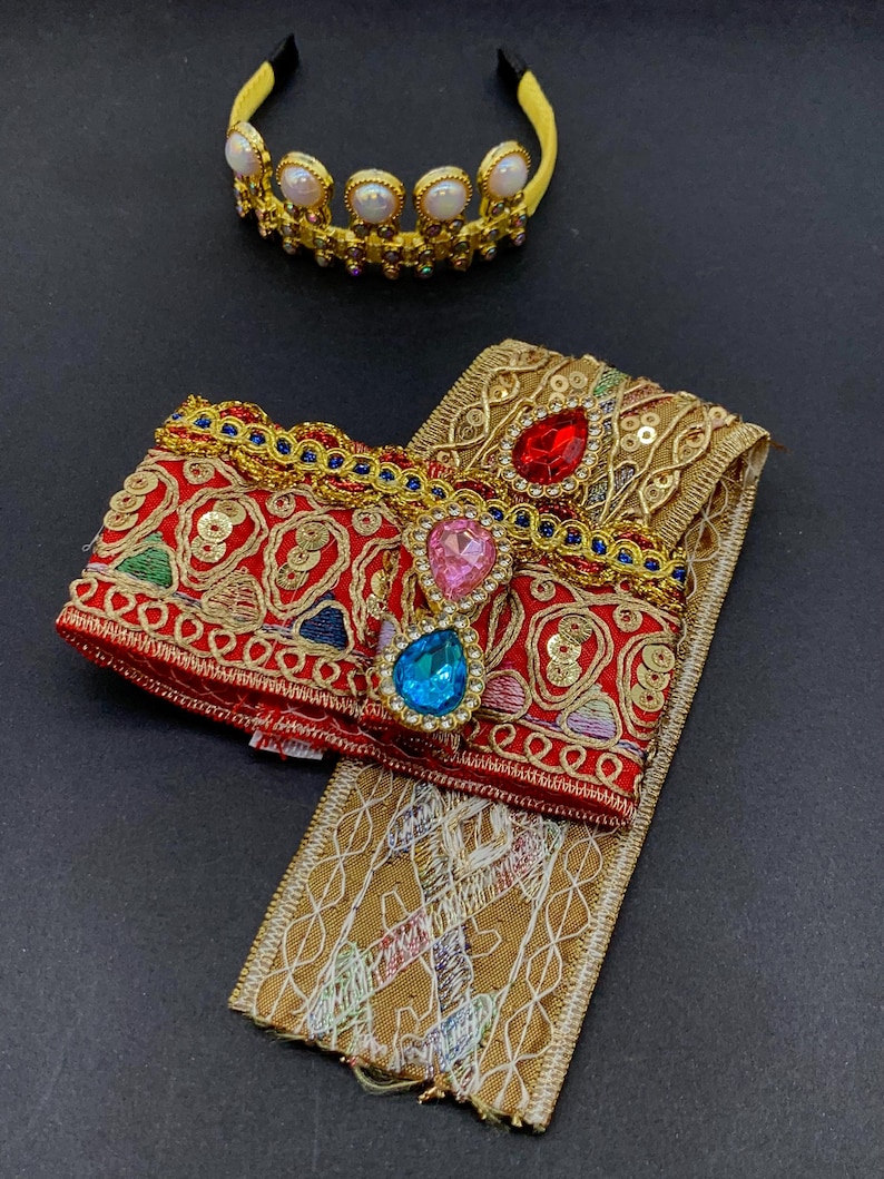 May include: A gold-colored crown with white pearl-like accents and a yellow headband. A red and gold embroidered accessory with colorful gems and a gold-colored trim. A gold-colored embroidered fabric.