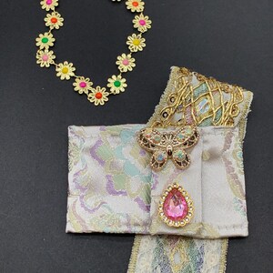 May include: A gold-toned daisy chain bracelet with colorful center accents. A decorative fabric piece with a butterfly brooch and a pink teardrop-shaped jewel. The fabric has a pastel pattern and gold trim.