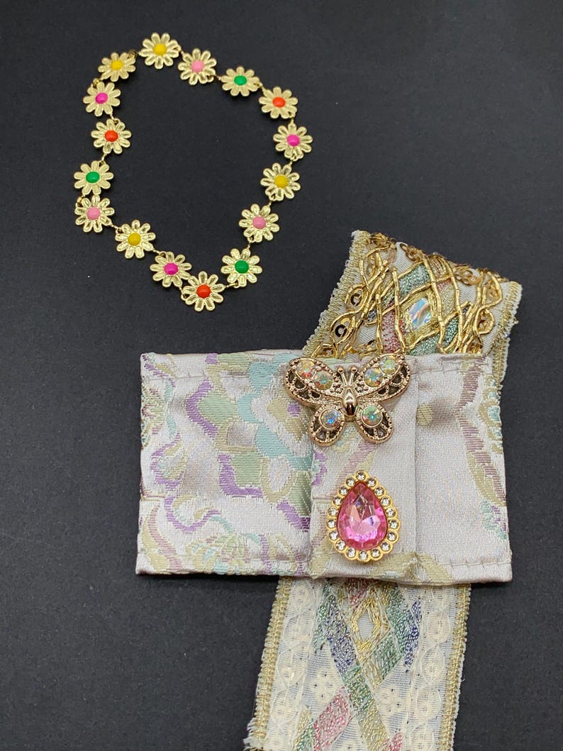 May include: A gold-toned daisy chain bracelet with colorful center accents. A decorative fabric piece with a butterfly brooch and a pink teardrop-shaped jewel. The fabric has a pastel pattern and gold trim.