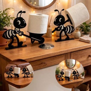 May include: Two black metal ant-shaped toilet paper holders. One ant figure pushes a wheelbarrow holding a roll of toilet paper, while the other ant figure stands upright and carries a roll of toilet paper on its back. Both figures have white accents.