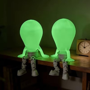 May include: Two green glowing light bulb figures with arms and legs, sitting on a wooden surface. The light bulbs have a bulb shape and are attached to a gray base. A small wooden clock is visible in the background.