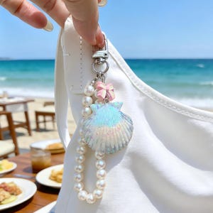 Peut inclure: Un porte-clés en forme de coquillage aux couleurs bleu et blanc irisé, orné d'un nœud rose et d'une rangée de fausses perles. Le porte-clés est attaché à un fermoir argenté. La scène de fond montre une plage.
