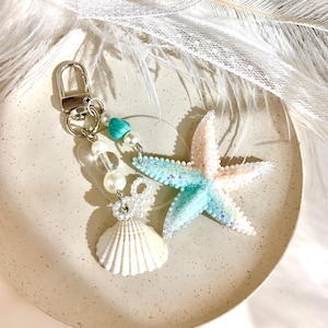 Peut inclure: Un porte-clés sur le thème de la plage avec un charme en forme d'étoile de mer et de coquillage. L'étoile de mer est bicolore, avec des sections rose clair et turquoise, et est ornée de paillettes. Le porte-clés comprend également un coquillage blanc, des perles et un fermoir argenté.