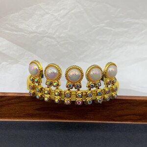 May include: A gold-toned tiara-style headband with iridescent pearl-like accents. The headband features a series of round, pearl-like stones set in gold frames, interspersed with smaller, colorful, iridescent stones. The design is symmetrical and ornate, suitable for special occasions.